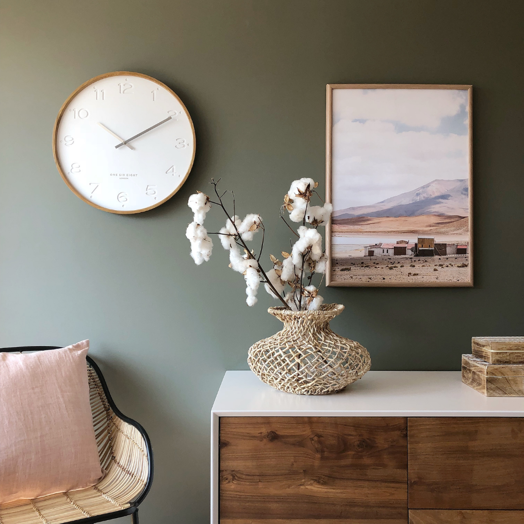 Modern home office scene featuring a white wall clock with gold accents mounted on an olive-green wall, styled with a rattan chair, blush cushion, cotton-stem vase, and framed landscape artwork.